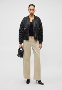 Γυναίκα που φοράει μαύρο bomber τζάκετ, μαύρο τοπ, μπεζ παντελόνι φαρδιάς γραμμής, μαύρα μποτάκια με τακούνι, κρατώντας μαύρη τσάντα με ασημένια τρουκς.