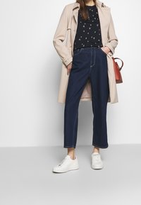 Imperméable beige, haut floral bleu marine, jean bleu foncé taille haute et baskets blanches. Les jeans ont des coutures visibles et une coupe ample.