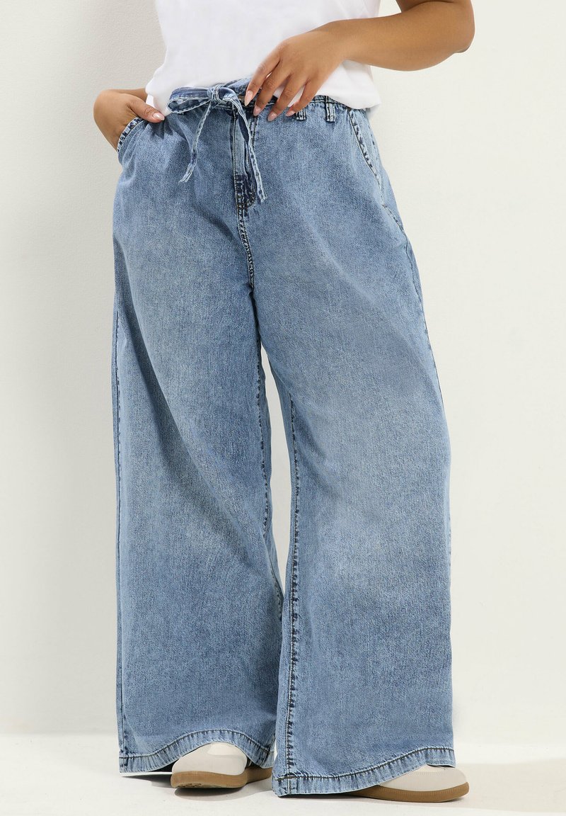 Person trägt hellblaue Jeans mit weitem Bein und gebundener Taille, weißes Oberteil und weiße Schuhe, steht mit Händen in den Taschen.