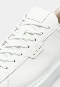 White leather sneaker featuring a stitched design, flat white laces, and a gold "NUBIKK" logo on the side. Smooth texture throughout.