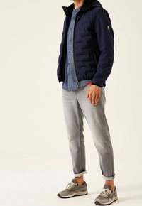 Man med marinblå vadderad jacka, blå knapp-up skjorta, ljusgrå uppvikta jeans och grå sneakers på ljus bakgrund.