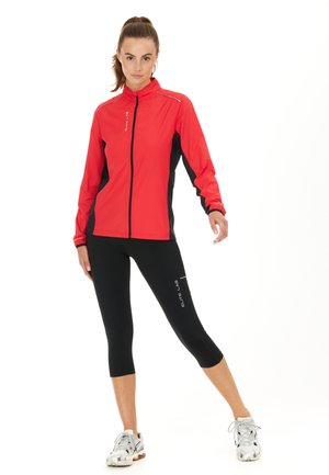 Rode sportjack met zwarte accenten, ritssluiting en reflecterende details; gecombineerd met zwarte cropped leggings en hardloopschoenen.