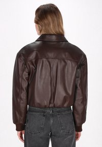 Bruine leren jas met een kraag, geribbelde manchetten en een tailleband. De achterkant heeft een centrale naad en een gladde textuur. Gekleed met donkerblauwe spijkerbroek.