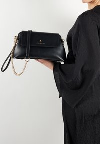 Borsa a tracolla nera in pelle con forma rettangolare, tracolla in catena dorata, chiusura a pattina e logo impresso sul davanti, tenuta in mano.