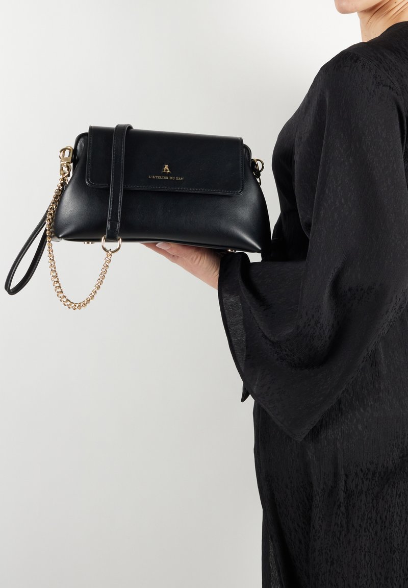 Borsa a tracolla nera in pelle con forma rettangolare, tracolla in catena dorata, chiusura a pattina e logo impresso sul davanti, tenuta in mano.