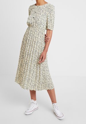 Vrouw die een lichtgele midi-jurk met een kleine bloemenprint draagt en witte hoge sneakers, staand tegen een effen lichte achtergrond.