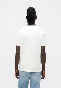 Bela bombažna majica kratkih rokavov z okroglim izrezom in sproščenim krojem, prikazana od zadaj, kombinirana s svetlo modrimi jeans hlačami.