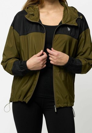 Personne portant une veste à capuche zippée vert olive et noir par-dessus un haut noir, les mains ajustent la fermeture éclair de la veste à l'avant.