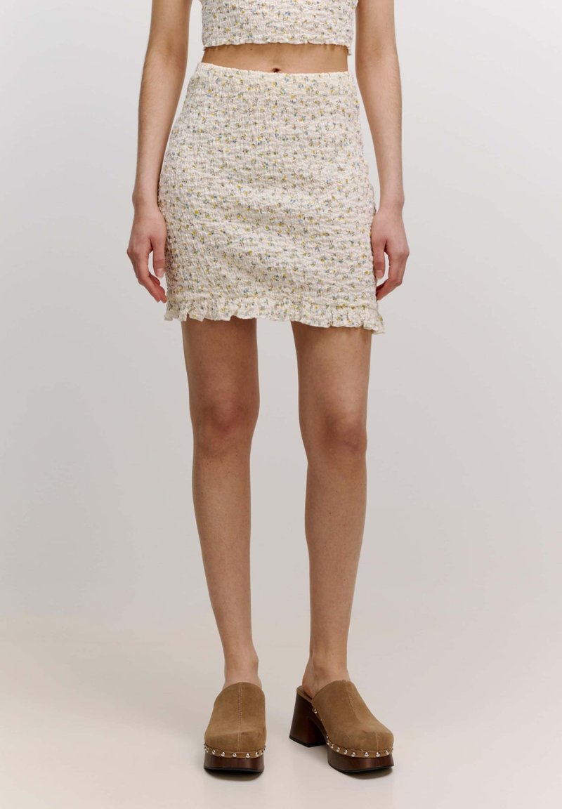 Jupe mini blanche avec un motif floral, en tissu texturé et à ourlet froncé. Associée à des mules en daim marron avec des clous décoratifs et un talon épais.