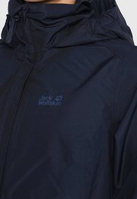 Marinblå vattentät jacka med huva, med en frontdragkedja och elastiska ärmslut. Logotyper är präglade på tyget. Slät textur.