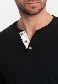 Hombre vistiendo una camiseta Henley negra con una tapeta blanca y tres botones en color marrón claro, mostrando el cuello y la parte inferior de la cara con barba recortada.