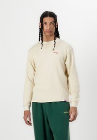 Beige long-sleeve top with ribbed texture, featuring a small red logo on the chest, paired with green sweatpants and a relaxed fit.