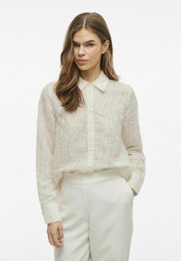 Camisa de botón de ajuste suelto en color crema, con una tela texturizada y suave, cuello clásico y mangas largas. Combinada con pantalones blancos.
