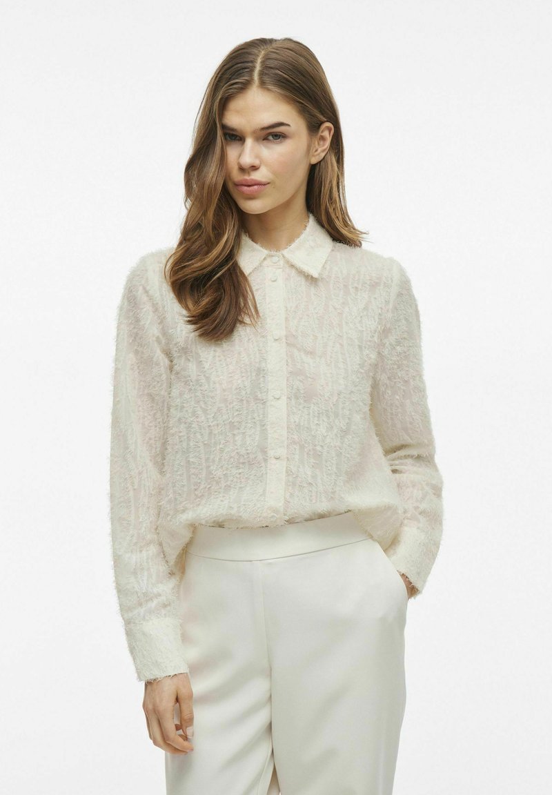 Camisa de botón de ajuste suelto en color crema, con una tela texturizada y suave, cuello clásico y mangas largas. Combinada con pantalones blancos.