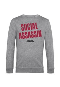Grå långärmad sweatshirt med djärv röd text som står "SOCIAL ASSASSIN" och "Curb Your Enthusiasm" i mindre svart text.