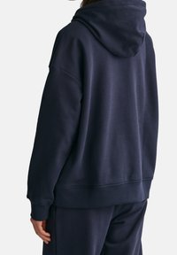 Persona che indossa un hoodie blu navy scuro e pantaloni coordinati, vista di spalle, che mostra i dettagli della parte posteriore e del cappuccio su uno sfondo semplice.