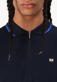 Polo shirt bleu marine avec un col zippé, des accents de col bleu contrastants, un tissu texturé et un petit patch logo sur l'avant.