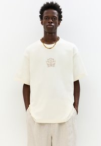 Cream oversized t-shirt with short sleeves, featuring a subtle embroidered floral design in brown at the center, worn with beige trousers.
