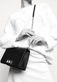 Model wears a white sweater and holds a black handbag with a chain strap, showcasing a stylish outfit in a monochromatic setting.