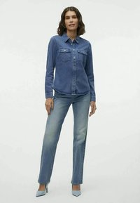Camisa de denim azul com bolsos frontais, design com botões, usada com jeans de denim azul claro e sapatos de salto alto azul pálido. Textura suave.