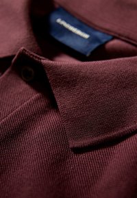 Närbild av en vikt, marinblå texturerad polo skjorta med krage och knapp, där tygweben syns och en del av en mörkblå etikett är synlig.