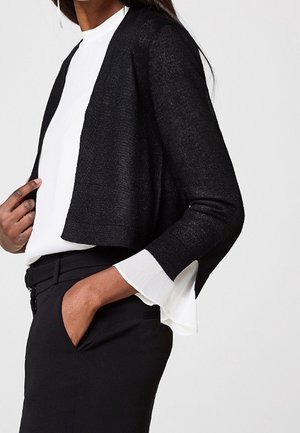 Kvinde iført en sort cropped cardigan over en hvid bluse med klokkeformede manchetter, kombineret med sorte højttaljede bukser.