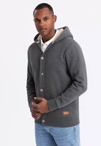 Grå hætte sweatshirt med knaplukning foran, der har en lys farvet foring, ribbede manchetter og en struktureret overflade. Lædermærke detalje på siden.