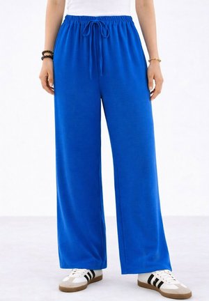 Personne portant un pantalon large bleu vif avec taille à cordon, des baskets blanches à rayures noires et un haut blanc, debout sur un sol clair.