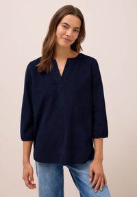 Vrouw met een lichte huid en bruin haar draagt een marineblauwe blouse met lange mouwen en lichtblauwe jeans, staande tegen een lichtbeige achtergrond.