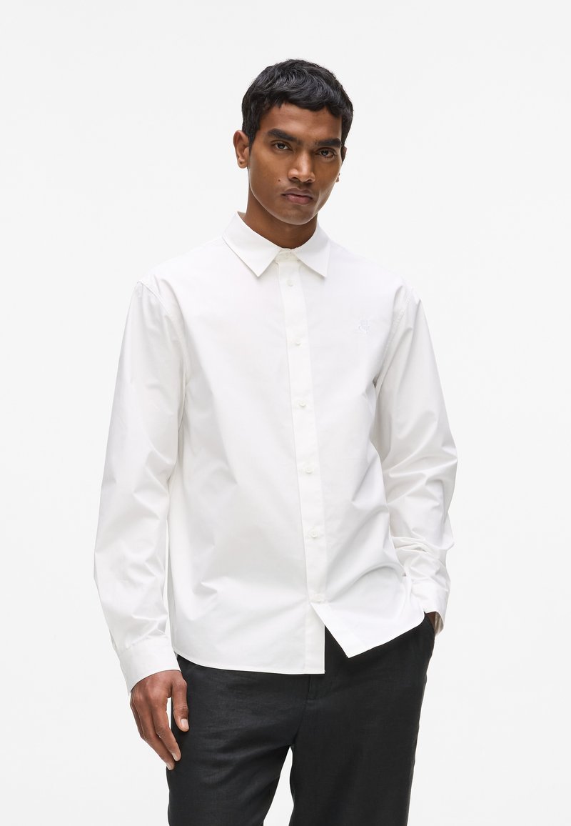 Chemise blanche à manches longues en coton, avec un design boutonné, un col pointu et un logo brodé discret sur le côté gauche de la poitrine.