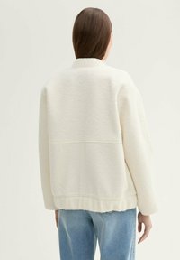 Weißer, strukturierter Fleece-Jacke mit lockerem Schnitt, rundem Halsausschnitt und geripptem Saum, kombiniert mit hellblauen Jeans. Dezente Nähte.