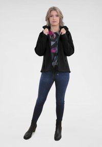 Zwarte zip-up jas met een hoge kraag, gedragen over een multicolored patroon top, gecombineerd met donkere skinny jeans en enkellaarzen.