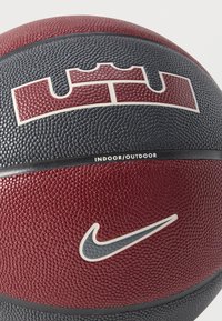 Minge de baschet cu suprafață texturată neagră și maro închis, având un logo distinctiv în formă de coroană și simbolul Nike Swoosh, etichetată pentru utilizare în interior/exterior.