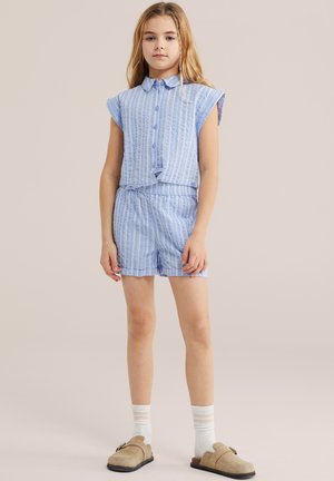 Junges Mädchen steht und trägt ein hellblau gestreiftes Hemd mit kurzen Ärmeln und passende Shorts, weiße Socken und beige Slipper.