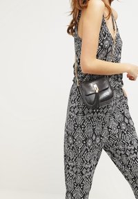 Mujer vestida con un mono de estampado geométrico en blanco y negro, llevando un pequeño bolso de cuero negro con cadena dorada y borlas.