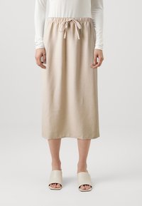 Jupe midi beige en tissu doux, dotée d'une taille élastique avec cordon de serrage et d'une coupe droite. Portée avec des claquettes de couleur claire.