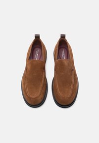 Brown suede slip-on shoes with a rounded toe, contrasting white stitching, and rubber sole. Features a pull tab for easy wear.