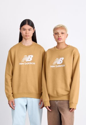 Braune Crew-Neck-Sweatshirts mit weißem "New Balance"-Logo, getragen mit hellblauen und braunen Hosen. Beide Sweatshirts verfügen über gerippte Bündchen und Säume.