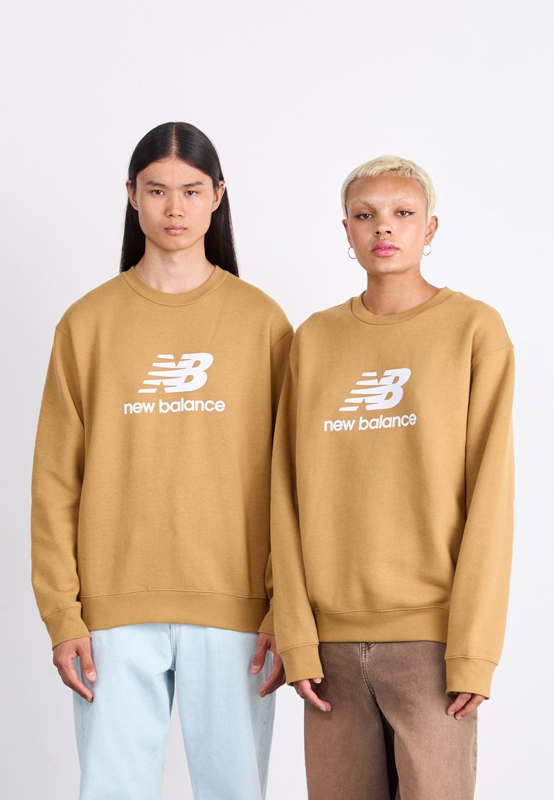 Smeđi dukserice s okruglim izrezom i belim logotipom "New Balance", nošene uz svijetloplave i smeđe hlače. Obje dukserice imaju rebraste manžete i donji rub.