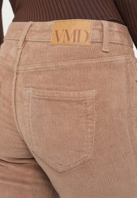 Pantalon en velours côtelé beige avec une poche arrière et un patch en cuir marqué VMD, porté avec un haut côtelé marron.