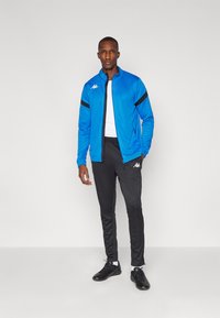 Veste de sport bleue avec accents noirs, logo blanc ; associée à un pantalon de survêtement noir ajusté et des chaussures de sport noires. Tissu lisse, zip intégral.