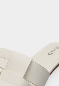 Gros plan sur une sandale beige sans lacets avec de larges brides chevauchantes et des coutures visibles près de la semelle.