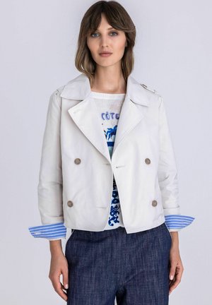 Femme portant une veste blanche à double boutonnage sur un pull à motifs, avec des manches de chemise bleue rayée retroussées et un pantalon foncé.