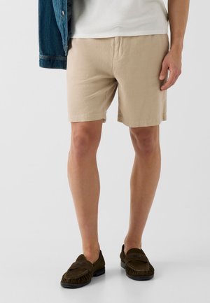 Man draagt beige korte broek, wit overhemd, donkerbruine loafers en houdt een spijkerjasje vast tegen een effen lichte achtergrond.