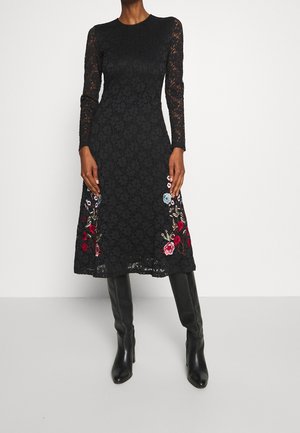 Woman wearing a black lace midi dress with long sleeves and floral embroidery near the hem, paired with knee-high black boots.