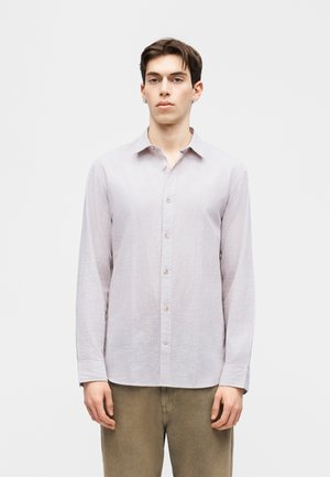 Νεαρός άνδρας όρθιος, φοράει ανοιχτόχρωμο ριγέ πουκάμισο με κουμπιά και μακριά μανίκια και ελιάς-πράσινο παντελόνι, ουδέτερο φόντο.