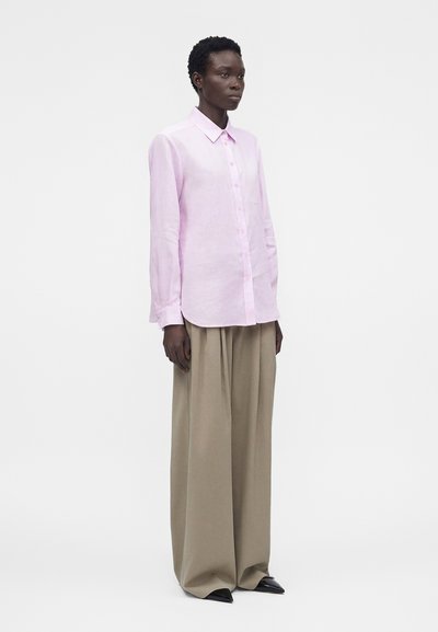 Μοντέλο φοράει ένα ανοιχτό ροζ πουκάμισο με μακριά μανίκια και κουμπιά, το οποίο είναι φορεμένο μέσα σε φαρδιά μπεζ παντελόνια με μαύρα μυτερά παπούτσια, στέκεται μπροστά σε απλό φόντο.