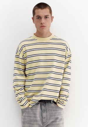 Ung mand med kort hår iført en gul langærmet skjorte med sorte striber og grå bukser, stående mod en hvid baggrund.