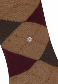 Brązowa, burgundowa i ciemnoszara tkanina w wzór argyle z pomarańczowymi przeszyciami oraz małym okrągłym metalowym emblematem na środku.