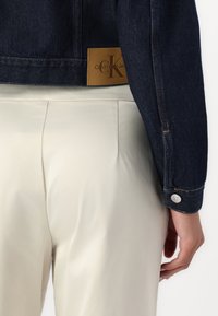 Tamno plavi traper jakna s smešnom kožnom oznakom na kojoj se nalazi logo "Calvin Klein Jeans". Svijetlosmeđe hlače su vidljive i prikazuju glatku teksturu.